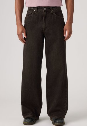 Personne portant un jean noir ample avec des poches avant, des chaussures noires et une chemise violet clair, debout devant un fond uni.