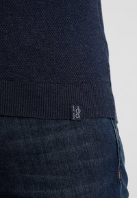Pull en maille bleu marine avec un motif texturé et un ourlet côtelé, arborant une étiquette sur le côté. Porté sur un jean en denim foncé.