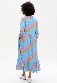 Žena s kudrnatými vlasy, oblečená v dlouhých šatech s potiskem srdcí v modré, růžové a hořčicové barvě, a bílých teniskách, stojící s rukama v kapsách, čelem odvrácena.