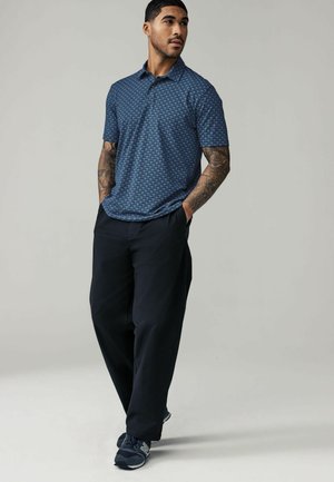 Blå kortærmet polo skjorte med et geometrisk mønster, parret med sorte løstsiddende bukser og mørke sneakers, båret af en tatueret mand.