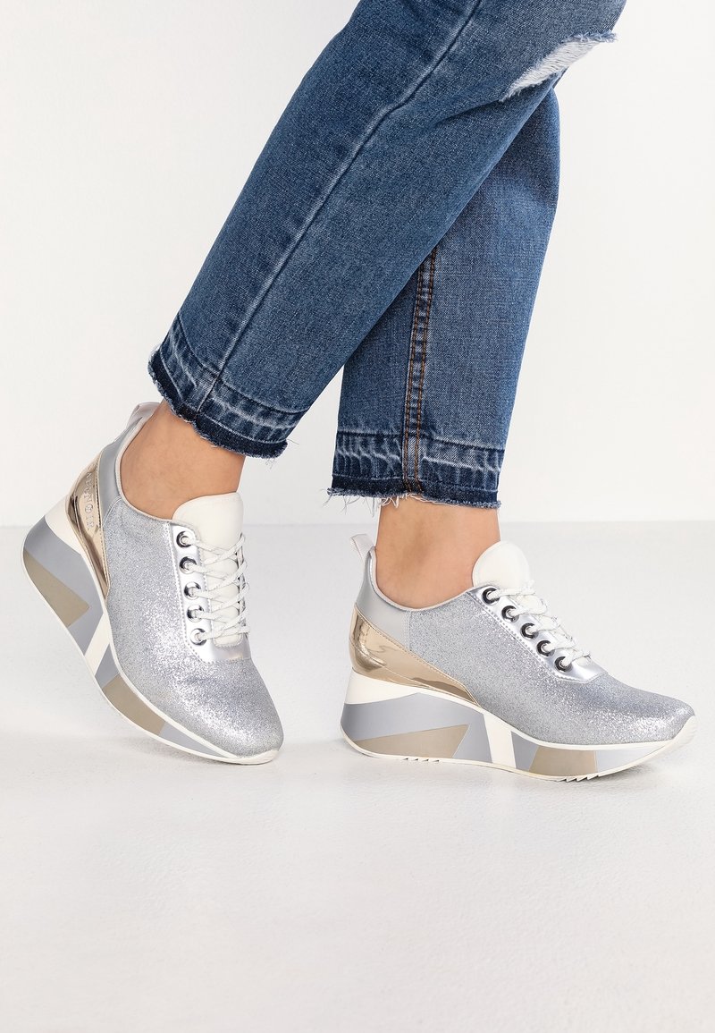 Jambes en jean denim bleu portant des baskets compensées argentées pailletées avec des lacets blancs et des accents dorés, debout sur un sol blanc.