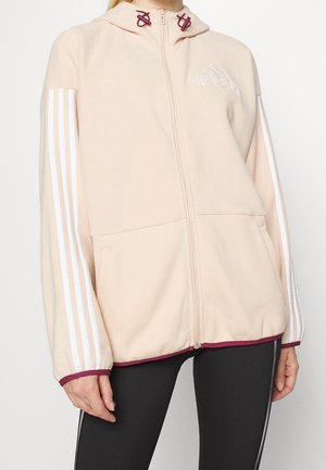 Femme portant un sweat à capuche zippé beige clair avec des rayures blanches sur les manches et des accents bordeaux, associé à des leggings noirs avec des coutures blanches sur les côtés.