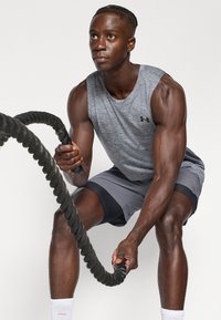 Szürke ujjatlan póló és szürke sportnadrág, vastag fekete hadit kötelet tartva. A háttér sima fehér. A fókusz a fitnesz tevékenységen van.