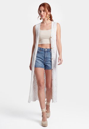 Kvinde med rødt hår iført en lang hvid hækle-vest, beige crop top, denimshorts, beige kilehælssko med ankelsnøre og runde øreringe, der går fremad.