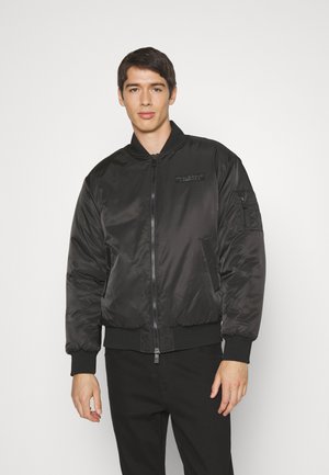Chaqueta bomber negra hecha de un material brillante y suave, con puños y dobladillo acanalados. Presenta un cierre frontal y un bolsillo con cremallera en la manga.