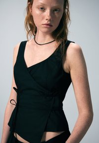 Haut noir sans manches avec un design asym étrique, un détail drapé et des liens latéraux. Texture lisse avec une ouverture fendue à l'ourlet.