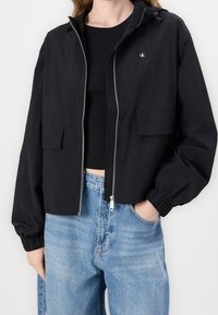 Czarna krótka kurtka z lekkiego materiału, z zamkiem z przodu, kapturem i dwiema przednimi kieszeniami. Rękawy z mankietami z elastycznymi detalami.