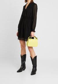 Schwarzes Hemdkleid mit transparenten langen Ärmeln und gerafftem Taillenbereich, kombiniert mit einer gelben Handtasche mit strukturiertem Finish und Kettenhenkel.