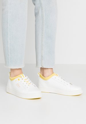 Voeten in witte sneakers met gele accenten en s.Oliver-logo, gecombineerd met lichtblauwe cropped jeans op een eenvoudige grijze achtergrond.