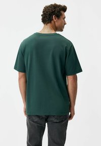 Zielona bawełniana koszulka z krótkimi rękawami, okrągłym dekoltem i prostym dołem, pokazana od tyłu na białym tle.