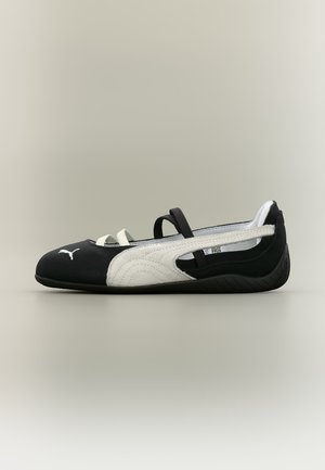Schwarzer und weißer Puma Ballerina-Schuh mit elastischen Riemen und Gummisohle, Seitenansicht vor schlichtem hellem Hintergrund.