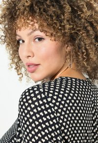 Haut à motifs noir et blanc avec de petites formes de cœur, doté d'un col arrondi et d'un tissu texturé. Le modèle a des cheveux bouclés et porte des boucles d'oreilles.