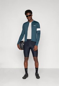 Férfi sötét teal kerékpáros dzsekit, fekete kerékpáros rövidnadrágot, napszemüveget és fekete cipőt visel, az egyik kezében egy fekete kerékpáros sisakot tart.