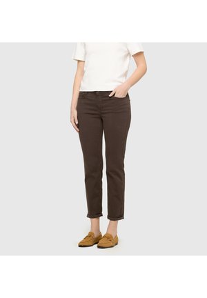 Persoon draagt donkerbruine rechte broek, wit T-shirt met korte mouwen en beige loafers, staand met één hand in de zak.
