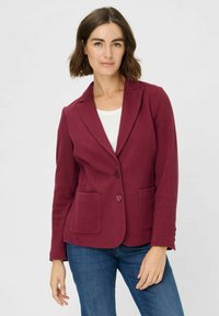 Bordeaux blazer van een textuurstof, met geknipte revers, twee voorzakken en een enkele knoopsluiting. Gecombineerd met een witte top en jeans.