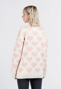 Maglione crema con grandi motivi a cuore rosa, texture lavorata a maglia e orlo a coste. Vestibilità oversize, indossato con jeans scuri.