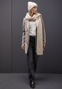 Beige wollen jas, lichtgrijze trui, zwarte jeans en een geribbelde crème sjaal en hoed. Zwarte enkellaarsjes maken de outfit compleet. Neutrale tinten.