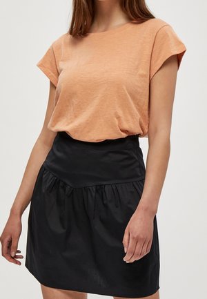 Femme portant un haut pêche à manches courtes rentré dans une jupe noire taille haute avec des fronces à la taille.