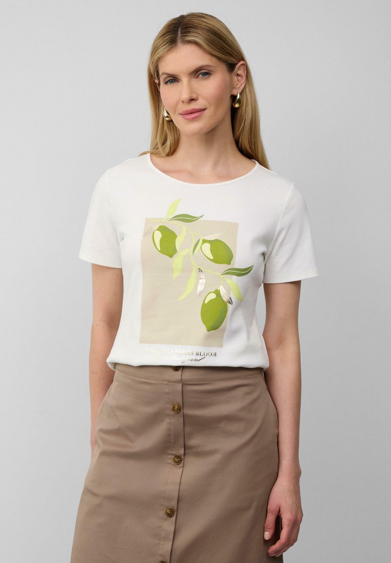 Witte t-shirt met korte mouwen, met een groene olijfafbeelding op een beige achtergrond. Gelaagd over een bruin, knoopsluiting rok.