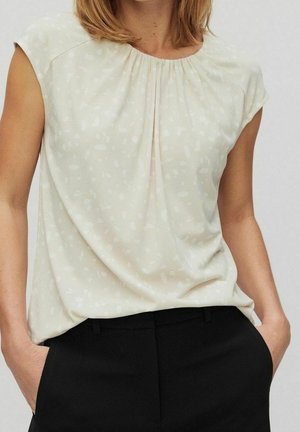 Mujer con blusa beige claro de manga corta con un patrón sutil y pantalones negros de cintura alta, con las manos en los bolsillos.