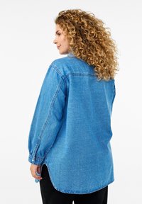 Camicia in denim di colore azzurro chiaro, con maniche lunghe, polsini abbottonati e orlo curvo. Il tessuto ha una consistenza liscia e cuciture visibili.