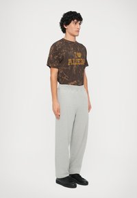 Camiseta marrón de manga corta con un estampado gráfico amarillo, combinada con pantalones de chándal grises y holgados. Zapatillas negras completan el conjunto.