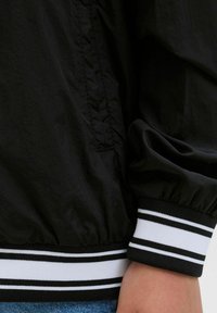 Juodas bombonešis su tekstūruotu paviršiumi. Yra elastingi rankogaliai su baltais ir juodais dryžiais. Matomas šoninis kišenės detalė.