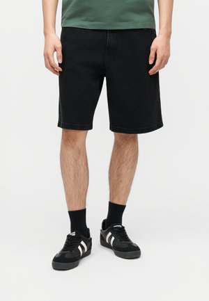 Mann trägt schwarze Jeansshorts, schwarze Socken und schwarze Sneakers mit weißen Streifen und steht vor einem schlichten weißen Hintergrund.