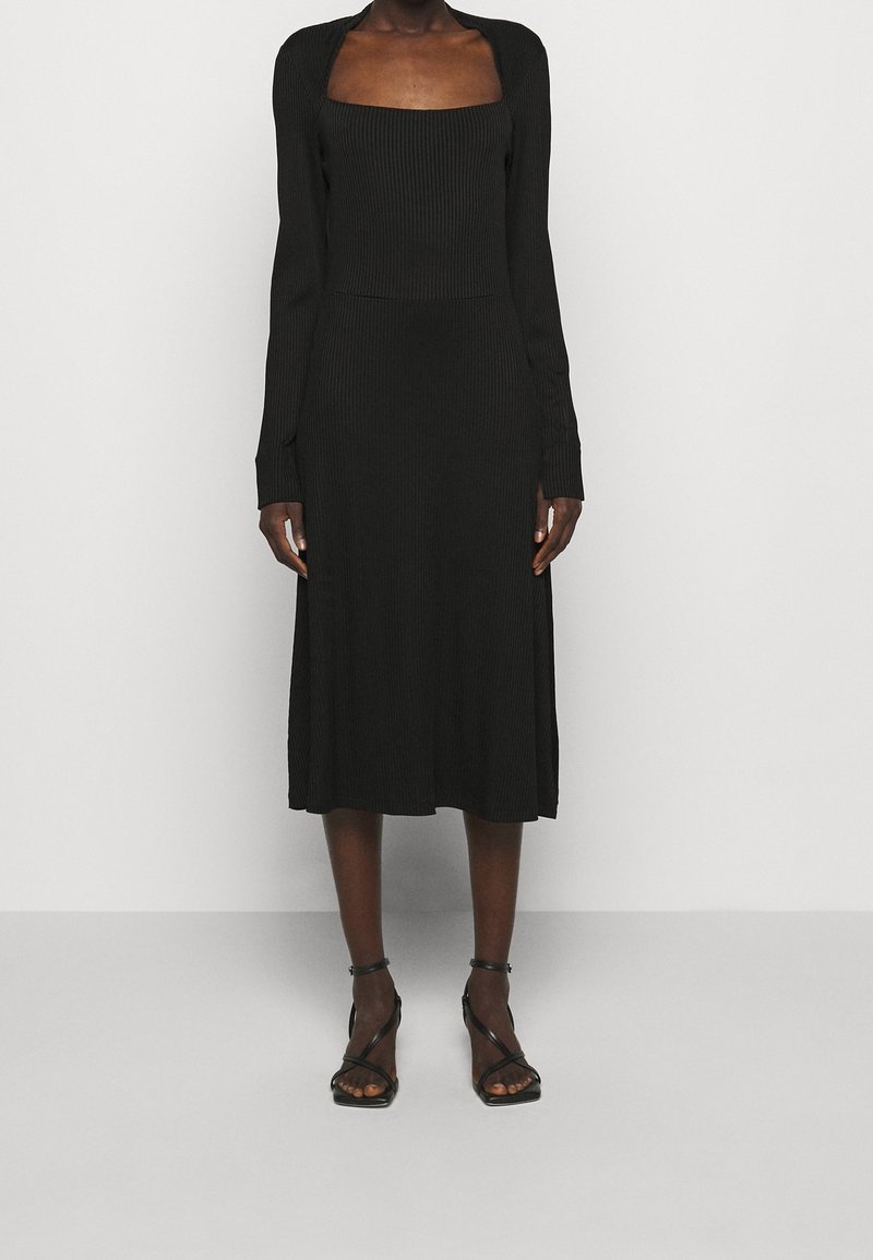 Černé proužkované midi šaty s dlouhými rukávy, čtvercovým výstřihem a těsným korzetem, který se na pase rozšiřuje. Nosené s černými sandály na podpatku.