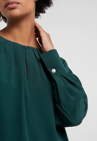Blusa verde con scollatura plissettata, tessuto morbido e maniche con bottone a perla. Texture liscia e vestibilità comoda.