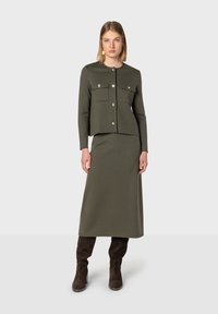 Giacca a maglia aderente verde oliva con bottoni argentati e due tasche sul petto, abbinata a una lunga gonna coordinata. Stivali marroni alti fino al ginocchio completano il look.