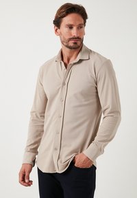 Camisa de manga larga color beige, hecha de una tela suave, con un cuello tipo solapa, cierre de botones en la parte delantera y mangas ajustadas. Textura sutil y aberturas laterales visibles.