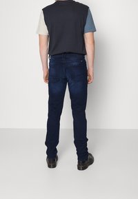 Vaqueros de hombre de color azul oscuro con un corte ajustado, que cuentan con dos bolsillos traseros y costuras sutiles. El modelo lleva una camiseta de manga corta con bloques de color.