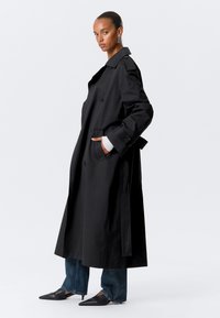 Cappotto lungo nero con una vestibilità ampia, design doppiopetto e tasche grandi. Indossato sopra jeans e scarpe a punta. Tessuto liscio.