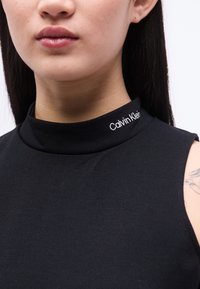 Γυναίκα που φοράει μαύρο τοπ χωρίς μανίκια με το λογότυπο "Calvin Klein" κεντημένο στον ψηλό γιακά, με μερικώς ορατό τατουάζ στον αριστερό ώμο.