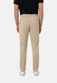 Beige Hose mit strukturierter Oberfläche, elastischem Bund, zwei Gesäßtaschen und schmal zulaufendem Design. Kombiniert mit weißen Sneakers.