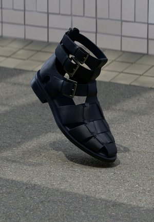 Schwarze Leder-Gladiator-Sandale mit drei verstellbaren Metallschnallen auf Fliesen- und Asphaltboden.