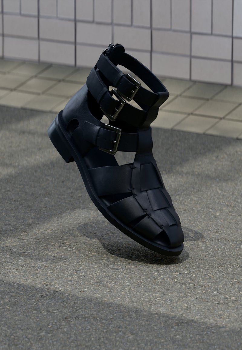 Μαύρο δερμάτινο σανδάλι γλαδιator με τρεις ρυθμιζόμενες μεταλλικές αγκράφες σε πλακωμένο και ασφαλτοστρωμένο έδαφος.