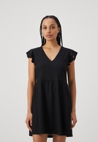 Vestido de algodão preto com decote em V, mangas curtas com babados e saia ampla. Apresenta padrões de ilhós por todo o vestido para adicionar textura.