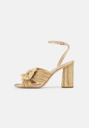 Gold heeled sandals with a knotted front, pleated texture, and an ankle strap. The block heel has a ribbed design, enhancing grip.