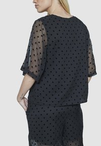 Kvindelig iført sort gennemsigtig top med prikker og matchende shorts, vist fra ryggen og siden, hånd i lommen.