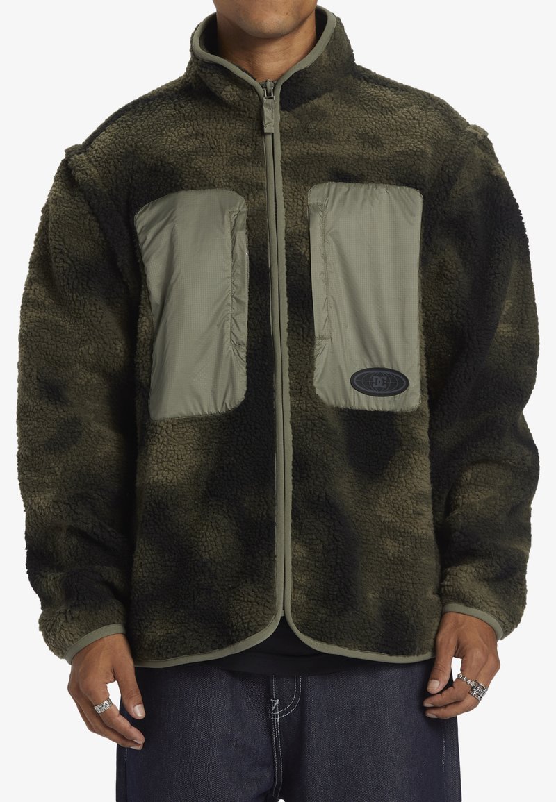 Chaqueta de forro polar en camuflaje verde oliva oscuro con cierre de cremallera, bolsillos en el pecho de nylon gris y cuello de canalé. Tejido texturizado y ajuste relajado.