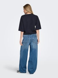 Ženska z dolgimi blond lasmi, oblečena v črno ohlapno majico in široke modre kavbojke, stoji obrnjena stran na enostavni belem ozadju.