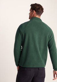 Green fleece pullover with a high collar and raglan sleeves. Soft texture, casual fit, paired with dark pants featuring back pockets.