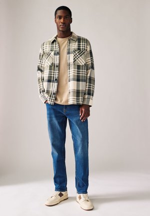 Bărbat purtând tricou bej, cămașă overshirt în carouri bej și negru, blugi albaștri și adidași bej, stând în fața unui fundal simplu, deschis.