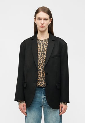 Vrouw met een oversized zwarte blazer over een trui met luipaardprint en blauwe spijkerbroek, staand tegen een witte achtergrond.