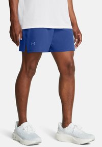 Bermudas deportivas azules con textura de malla, aberturas laterales y logo de Under Armour. Combinadas con zapatillas blancas que presentan una parte superior texturizada.