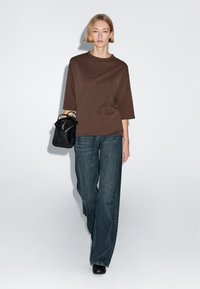 Woman in brown oversized t-shirt, wide-leg blue jeans, black shoes, holding small black handbag walking against white background.