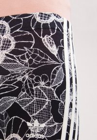 Mustia legginsit, joissa on valkoinen kukkakuvioinen pitsimalli, valkoiset sivu raidat ja Adidas-logo lähellä alaosaa. Joustava kangas.