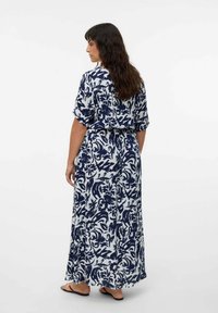 Donna con lunghi capelli scuri che indossa un maxi abito astratto blu navy e bianco, insieme a sandali neri piatti, in piedi su uno sfondo bianco semplice.